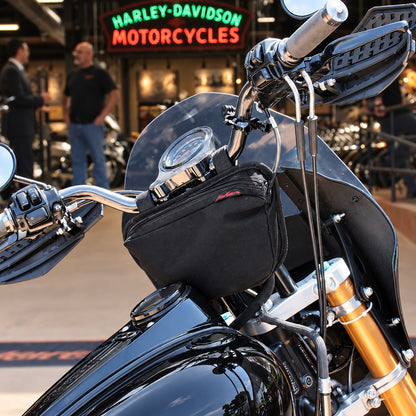 Black Moto Pockets OG T-Bar motorcycle handlebar bag mounted on a Harley-Davidson, showing its compact tapered design beneath the gauges with a Harley-Davidson dealership in the background.